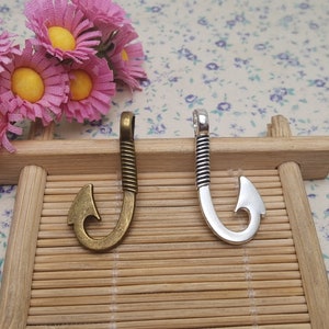 May include: Two metal fishing hook charms, one in antique brass and one in silver, on a wooden background.
