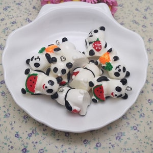 May include: A group of white panda bear charms with black eyes and black markings. Each charm has a different design, including a watermelon, a strawberry, and a carrot. The charms are on a white plate with a floral pattern.