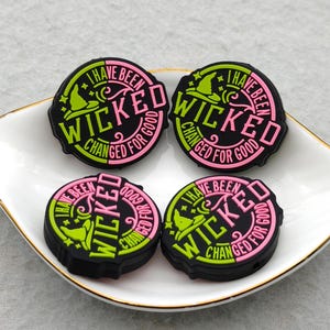 May include: Four black, circular charms with the text "I HAVE BEEN WICKED CHANGED FOR GOOD" in pink and green. Each charm features a witch hat and star design. The charms are displayed on a white, gold-rimmed dish.