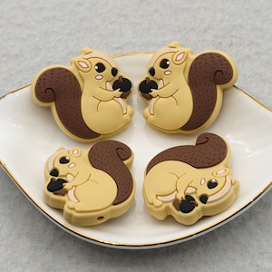 May include: Four squirrel-shaped embellishments in tan, brown, and black, arranged on a white, leaf-shaped dish with a gold rim. The squirrels are holding acorns, perfect for crafting or decorative projects.