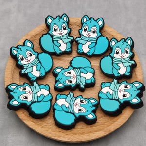 May include: A collection of teal squirrel-shaped embellishments with white accents and black outlines, arranged on a wooden plate. Each squirrel wears a white scarf. The plate sits on a gray surface.