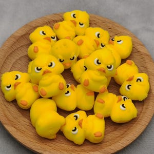 May include: A group of yellow rubber chick charms with black eyes and orange beaks. The charms are arranged in a circle on a wooden surface.