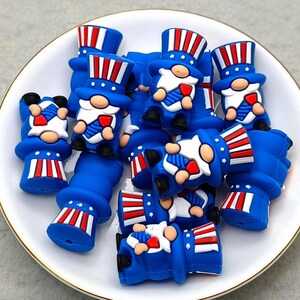 May include: A collection of blue gnome-shaped charms with red, white, and blue patriotic designs, including top hats and bow ties. The charms are arranged on a white plate with a gold rim, perfect for crafting or jewelry making.