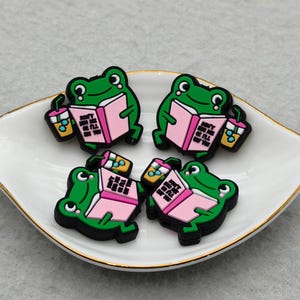 Op de afbeelding: Vier groene kikker-vormige bedels met zwarte contouren, elk met een roze boek en een drankje. De bedels zijn gerangschikt op een witte, bladvormige schaal met een gouden rand. De kikkers lezen boeken.