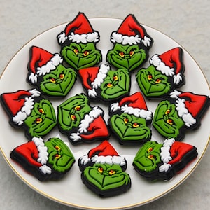 May include: A white plate with a gold rim displays several green, cartoon faces wearing red Santa hats with white trim. The faces have a grumpy expression. The plate is on a light grey surface.
