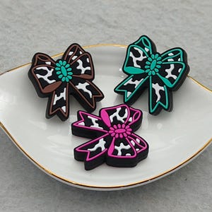 Op de afbeelding: Drie decoratieve strikvormige bedels in bruin, groen en roze, elk met een koeienprintpatroon. De strikken zijn gerangschikt op een wit, bladvormig schaaltje met een gouden rand. De bedels hebben een zwarte omtrek en een turquoise centrum.