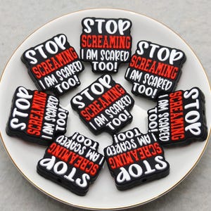 May include: A collection of black and white rectangular charms with the text "STOP SCREAMING I AM SCARED TOO!" in red and white lettering. The charms are arranged on a white plate with a gold rim.