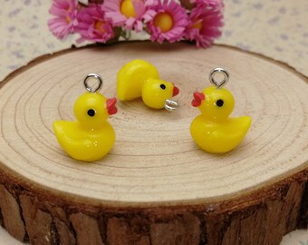 May include: Three yellow rubber duck charms with red beaks and black eyes. The charms are on a wooden surface with a pink flower in the background.