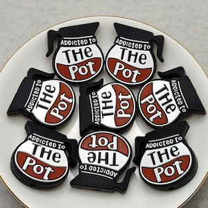 Può includere: Una collezione di ciondoli a forma di caffettiera neri, bianchi e rossi disposti su un piatto bianco con bordo dorato. Ogni ciondolo presenta la scritta "Addicted to The Pot".