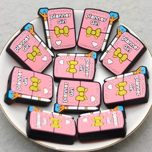 May include: A collection of pink, black, and yellow planner charms arranged on a white plate. Each charm features the text "planner Girl" and a yellow bow design. The charms are likely for crafting or decorating.