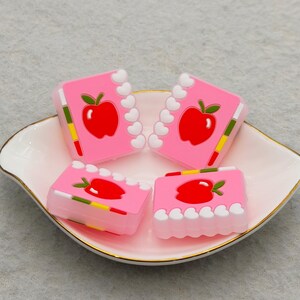 May include: Four pink, rectangular charms with a red apple design and white heart-shaped borders are arranged in a white and gold-rimmed dish. The charms have a notebook-like appearance, with green, yellow, and red accents.