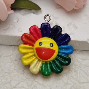 Puede incluir: Un amuleto de flor de colores arcoíris con una cara amarilla sonriente en el centro. El amuleto tiene un lazo plateado para sujetarlo a un collar o pulsera.