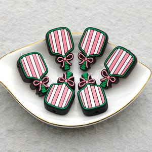 May include: Six decorative charms shaped like striped pink and white bottles with green and pink bows. The charms have a black border and are arranged on a white dish with a gold rim.