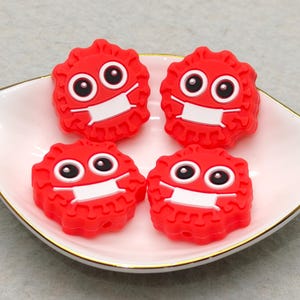 Puede incluir: Cuatro figuras rojas, parecidas a dibujos animados, con mascarillas blancas y grandes ojos negros, dispuestas sobre un plato blanco festoneado con borde dorado. Las figuras tienen forma circular y puntiaguda.