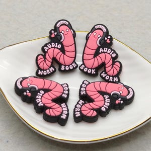 May include: Four pink and black shoe charms shaped like cartoon worms wearing headphones. Each charm has the words "Audio Book Worm" in white. The charms are arranged on a white, gold-rimmed plate.