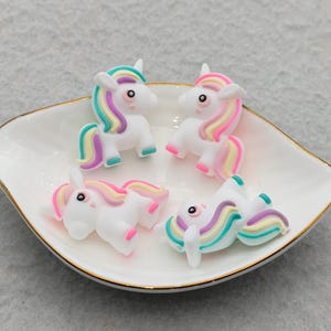 May include: Four small white unicorn figurines with multicolored manes and tails in shades of pink, yellow, blue, and purple. Each unicorn has a smooth finish, black eyes, and a horn on its forehead. The figurines are arranged on a white dish.