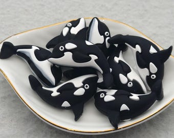 May include: A selection of black and white orca-shaped charms are displayed in a white dish with a gold rim. The orcas feature white markings and small, round eyes. The dish is on a light grey background.