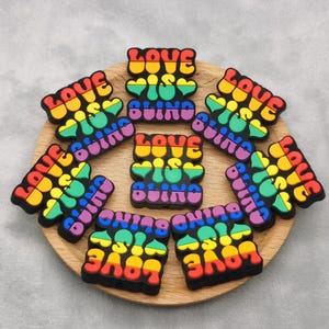 May include: A collection of colorful, rubbery shoe charms arranged on a wooden plate. Each charm features the phrase "LOVE IS BLIND" in a rainbow of colors, with a black border. The charms are a fun accessory.