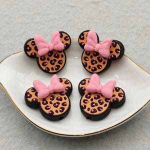 May include: Four decorative Minnie Mouse-shaped embellishments. Each has a black outline, a tan leopard print center, and a pink bow. They are arranged on a white, leaf-shaped dish with a gold rim.