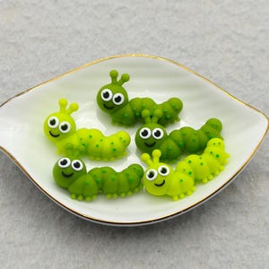 May include: A white, leaf-shaped dish with a gold rim holds six cartoon caterpillars. The caterpillars are various shades of green and yellow, with smiling faces and antennae. The dish sits on a light gray surface.