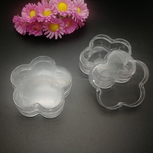 Op de afbeelding: Doorzichtige, bloemvormige plastic containers, gestapeld in groepen van drie. De containers staan op een zwart oppervlak, met een boeket roze en gele bloemen op de achtergrond.