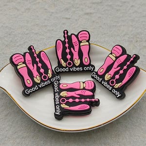 May include: A white ceramic dish with a gold rim holds four black and pink charms. Each charm features a pink design with the text "Good vibes only" in white. The charms are likely shoe decorations.