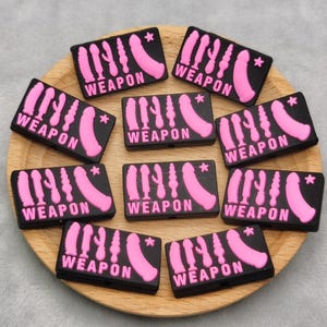 May include: A collection of black rectangular patches with the word "WEAPON" in white, and pink illustrations of sex toys. The patches are arranged on a wooden plate, with a gray background.