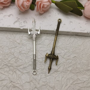 Pode incluir: Dois pingentes de tridente prateados, um com um design mais ornamentado, o outro com um design mais simples. Ambos os pingentes têm um laço na parte superior para serem presos a joias ou outros trabalhos manuais.