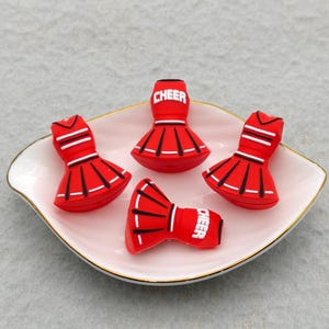Puede incluir: Cuatro dijes de falda de animadora en rojo y blanco con rayas verticales negras y la palabra CHEER impresa en blanco en la cintura. Estos pequeños accesorios moldeados tienen una forma acampanada y están dispuestos en un plato rosa pálido.