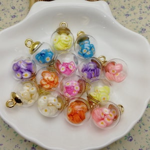 Könnte beinhalten: Eine Sammlung von 12 kleinen Glasflaschen mit goldenen Verschlüssen, jede gefüllt mit bunten, blumenförmigen Charms. Die Flaschen sind in einer weißen Schüssel angeordnet.