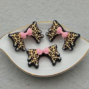 Op de afbeelding: Vier zwarte en goudkleurige schoenbedels met luipaardprint en roze strikken, gerangschikt op een wit schaaltje met een gouden rand. De bedels hebben een strikvorm en zijn versierd met kleine witte stippen.