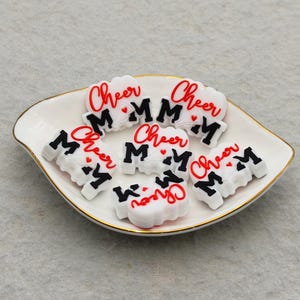 Puede incluir: Un plato blanco en forma de hoja con borde dorado contiene varios pequeños amuletos blancos en forma de nube. Cada amuleto tiene la palabra "Cheer" en escritura roja y las letras "M" en negro.
