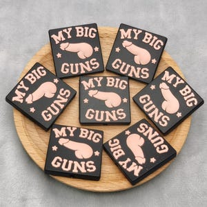 May include: A collection of black square magnets with the phrase "MY BIG GUNS" and a graphic of a penis in pink. The magnets are arranged on a round wooden tray, set against a gray background.
