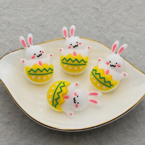 May include: A white ceramic dish with a gold rim holds four Easter-themed bunny figurines. The bunnies are white with pink ears and are emerging from yellow and green eggshell designs. The figurines are whimsical and decorative.