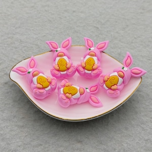 May include: Five pink bunny figurines with yellow egg accents are arranged on a pink, leaf-shaped dish with a gold rim. The bunnies have white floral accents and the word "hi" on the eggs. The dish sits on a light gray surface.