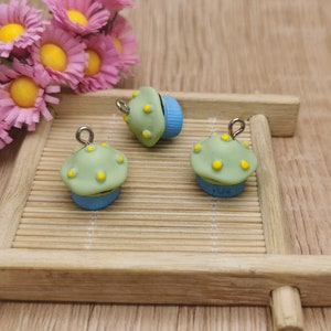 May include: Three green and blue cupcake charms with yellow sprinkles and silver jump rings. The charms are on a wooden surface.