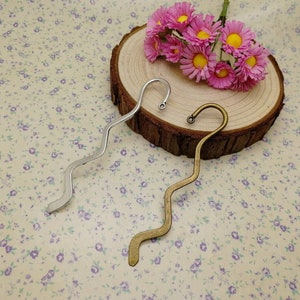 May include: Two silver and gold wavy bookmark with a hook at the top. The bookmarks are on a wooden surface with pink flowers.