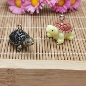 Puede incluir: Dos pequeños amuletos de tortuga, uno es negro y verde, el otro es amarillo con una concha marrón. Ambos tienen anillos de salto plateados.