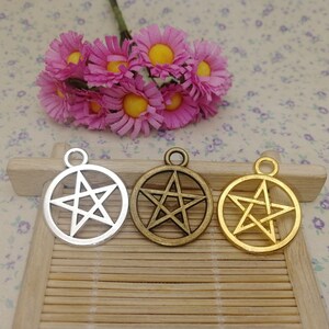 Puede incluir: Tres dijes de pentagrama plateados y dorados con un diseño de estrella de cinco puntas. Los dijes están sobre una superficie de madera.