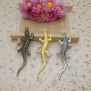 Puede incluir: Tres abalorios de metal con forma de lagarto en diferentes acabados: dorado, plateado y bronce. Cada abalorio presenta un diseño de lagarto detallado con escamas texturizadas.