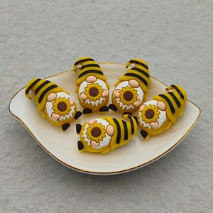 May include: A collection of five gnome figurines with yellow and black striped hats and bodies, each holding a sunflower. The gnomes are arranged on a white, scalloped-edge plate with a gold trim.
