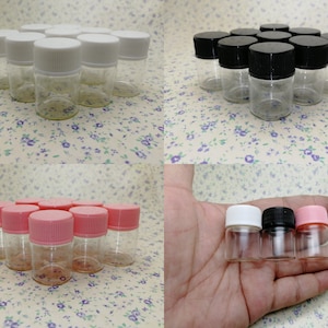 Op de afbeelding: Een set van heldere glazen flessen met witte, zwarte en roze deksels. De flessen zijn klein en rond en zijn gerangschikt in groepen van drie. De flessen zijn perfect voor het bewaren van kleine items, zoals etherische oliën, specerijen of sieraden.