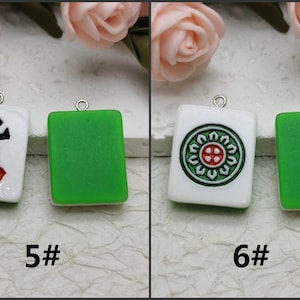 Puede incluir: Dos fichas de mahjong de cer&aacute;mica blanca con caracteres negros y rojos y un dise&ntilde;o de c&iacute;rculo verde. Una ficha tiene los caracteres chinos para "Nueve bamb&uacute;" y la otra tiene un c&iacute;rculo verde con una cruz roja en el centro. Ambas fichas tienen un lazo plateado para colgar.