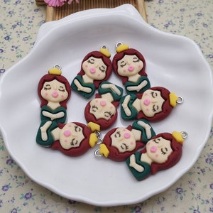 May include: A group of eight small, green and red clay charms shaped like girls with yellow crowns. Each charm has a silver loop for attaching to a necklace or bracelet.