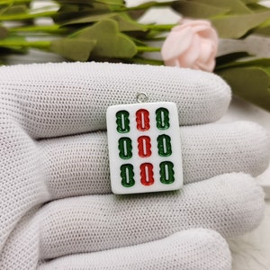 Puede incluir: Pendiente de cer&aacute;mica blanca en forma de ficha de mahjong con caracteres verdes y rojos. La ficha es un amuleto con un lazo plateado para colgar.