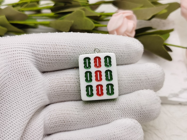 Puede incluir: Pendiente de cer&aacute;mica blanca en forma de ficha de mahjong con caracteres verdes y rojos. La ficha es un amuleto con un lazo plateado para colgar.