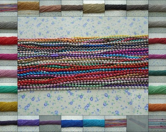 Può includere: Una collezione di maglie di catena metalliche colorate in vari colori, tra cui argento, oro, rosso, blu, verde e viola. Le catene sono disposte in file su uno sfondo bianco con un motivo floreale.