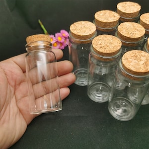 Puede incluir: Un grupo de botellas de vidrio transparente con tapones de corcho. Las botellas están dispuestas en semicírculo sobre una superficie negra. Una botella está siendo sostenida en una mano.
