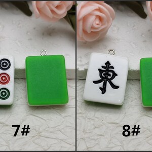 Puede incluir: Dos azulejos de cer&aacute;mica blanca con dise&ntilde;os negros, uno con tres c&iacute;rculos y otro con el car&aacute;cter chino "Este", colgando de cadenas plateadas. Los azulejos est&aacute;n sobre un fondo blanco.