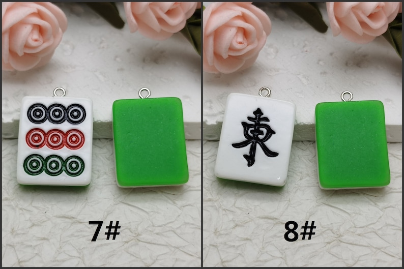 Puede incluir: Dos azulejos de cer&aacute;mica blanca con dise&ntilde;os negros, uno con tres c&iacute;rculos y otro con el car&aacute;cter chino "Este", colgando de cadenas plateadas. Los azulejos est&aacute;n sobre un fondo blanco.